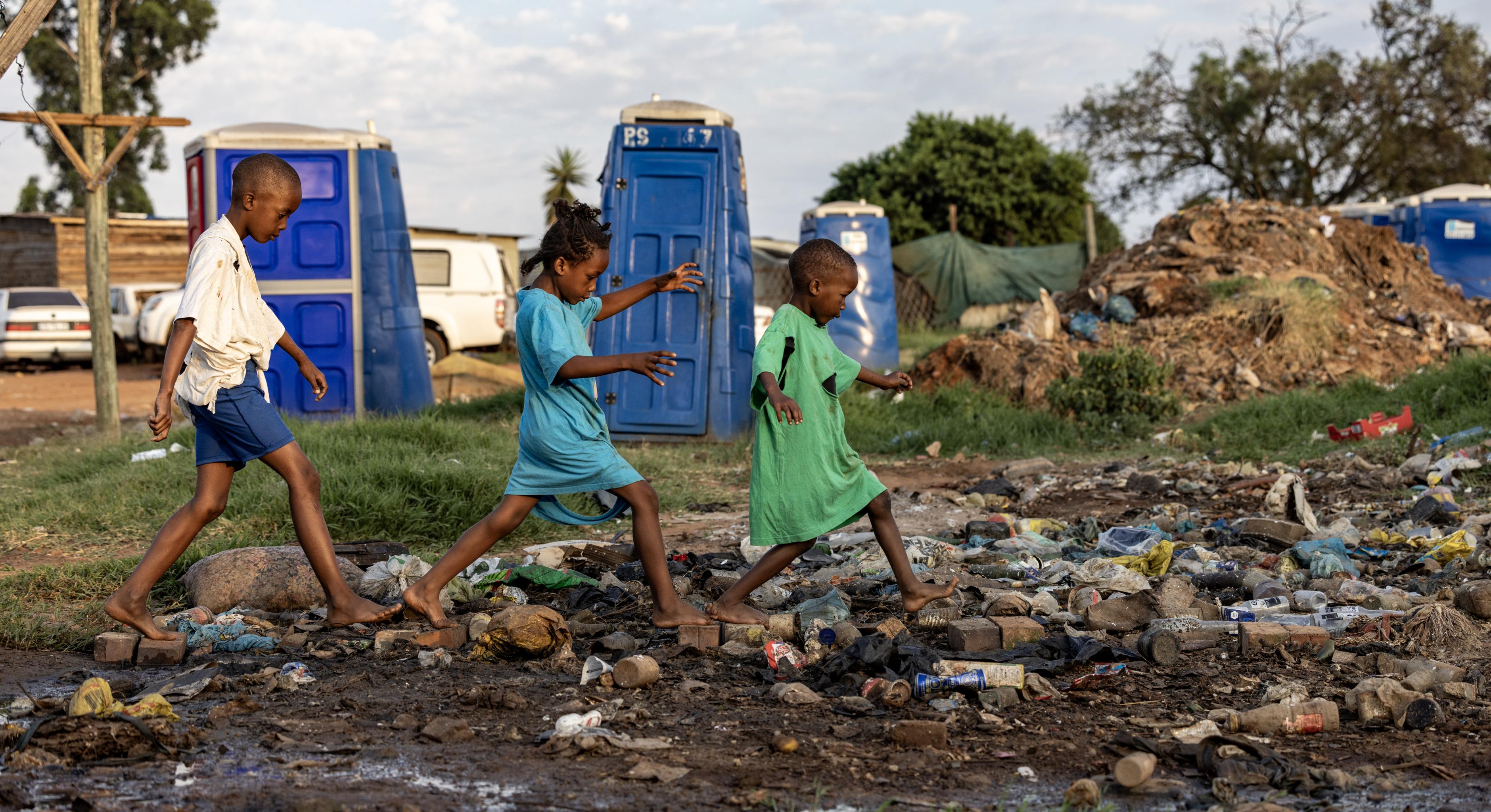 Drei Kinder laufen über einen Weg aus Steinen, Die Umgebung sieht sehr schmutzig aus. Müll liegt auf dem Weg. Abwasser scheint aus nahegelegenen Toiletten auszutreten.