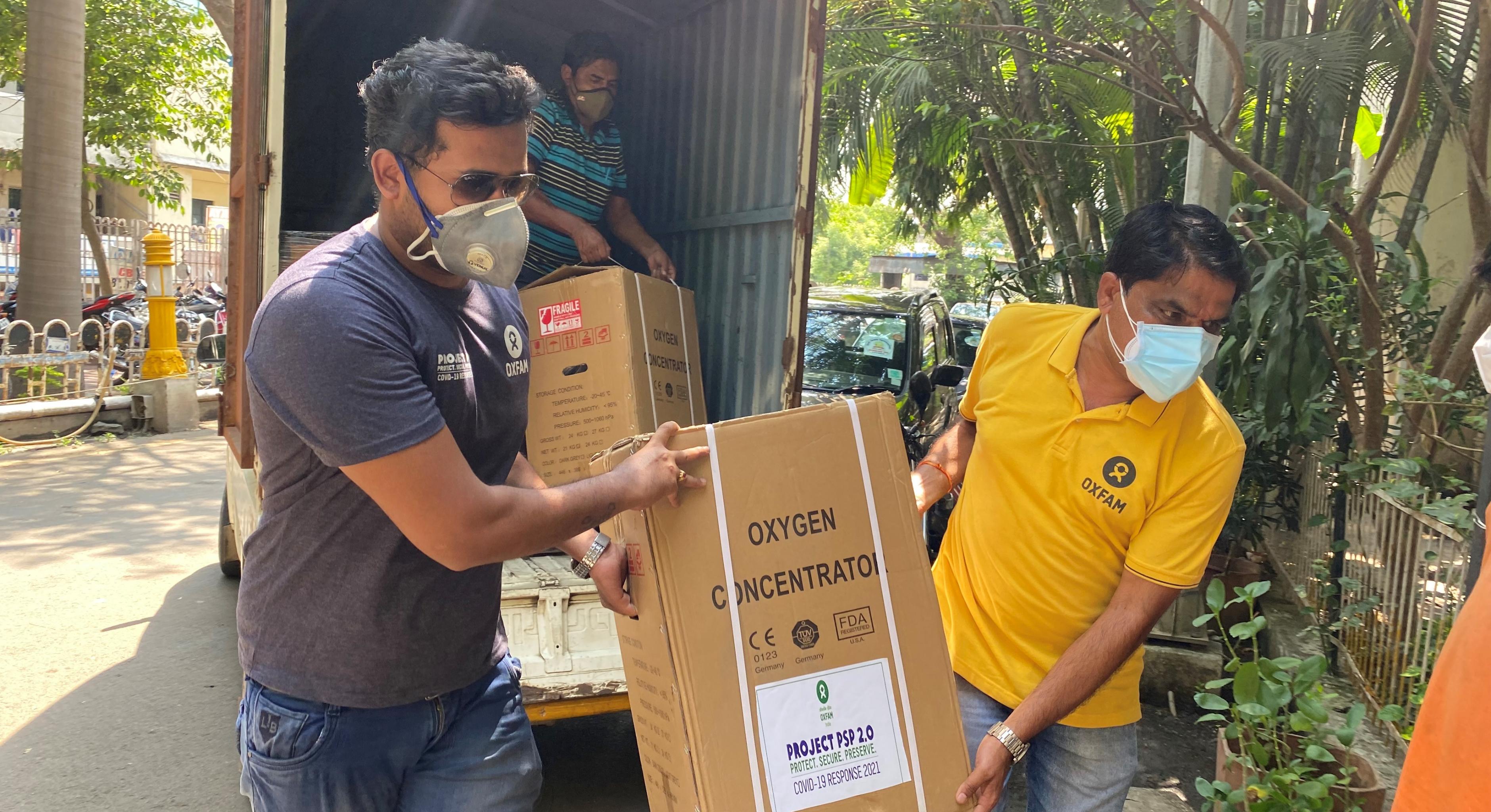 Zwei Mitarbeiter von Oxfam Indien tragen einen Karton mit einem Sauerstoffkonzentrator.