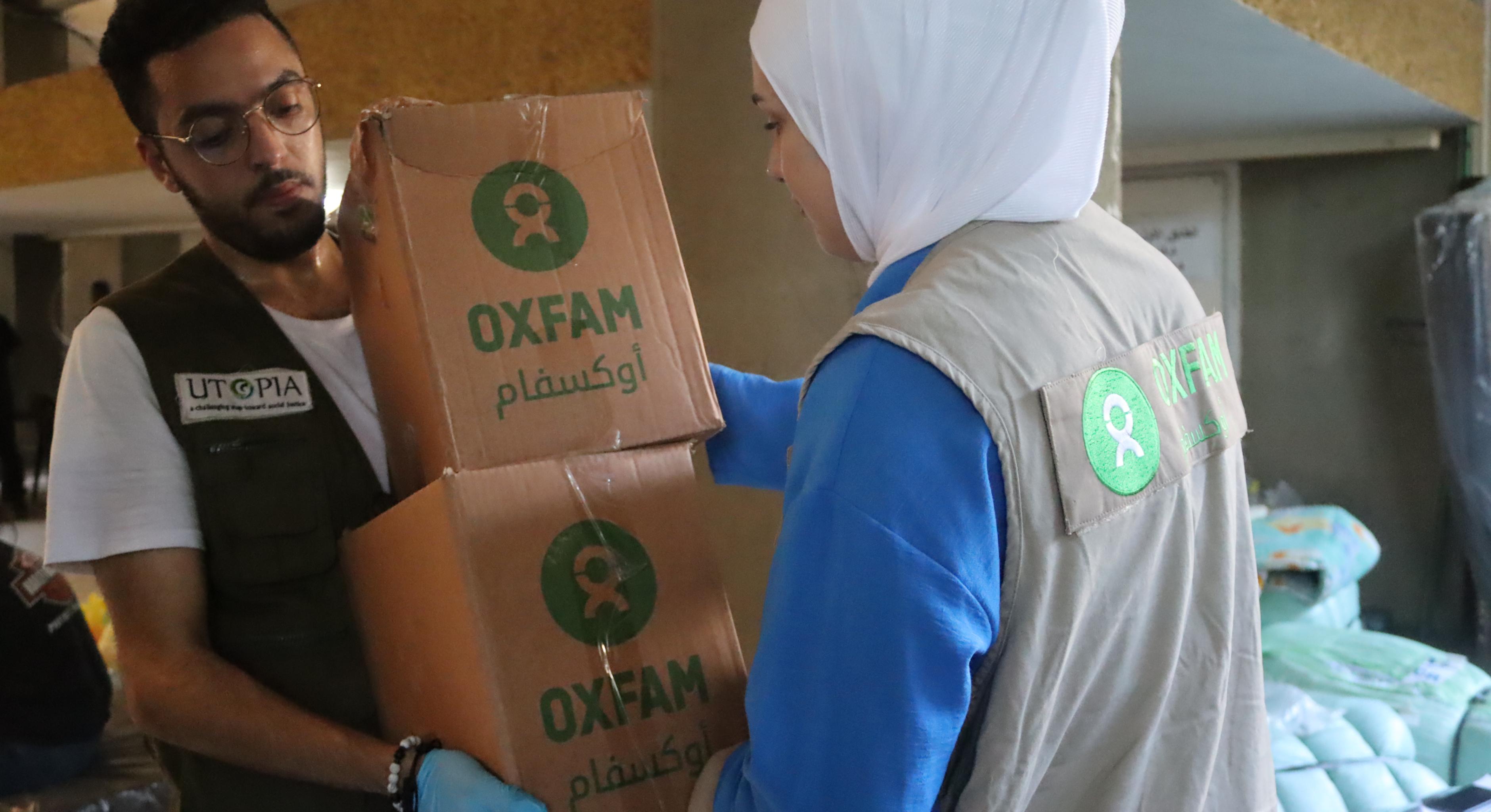 Eine Person übergibt zwei Kartons mit Oxfam-Aufschrift an eine zweite Person. Im Hintergrund stehen Feldbetten.