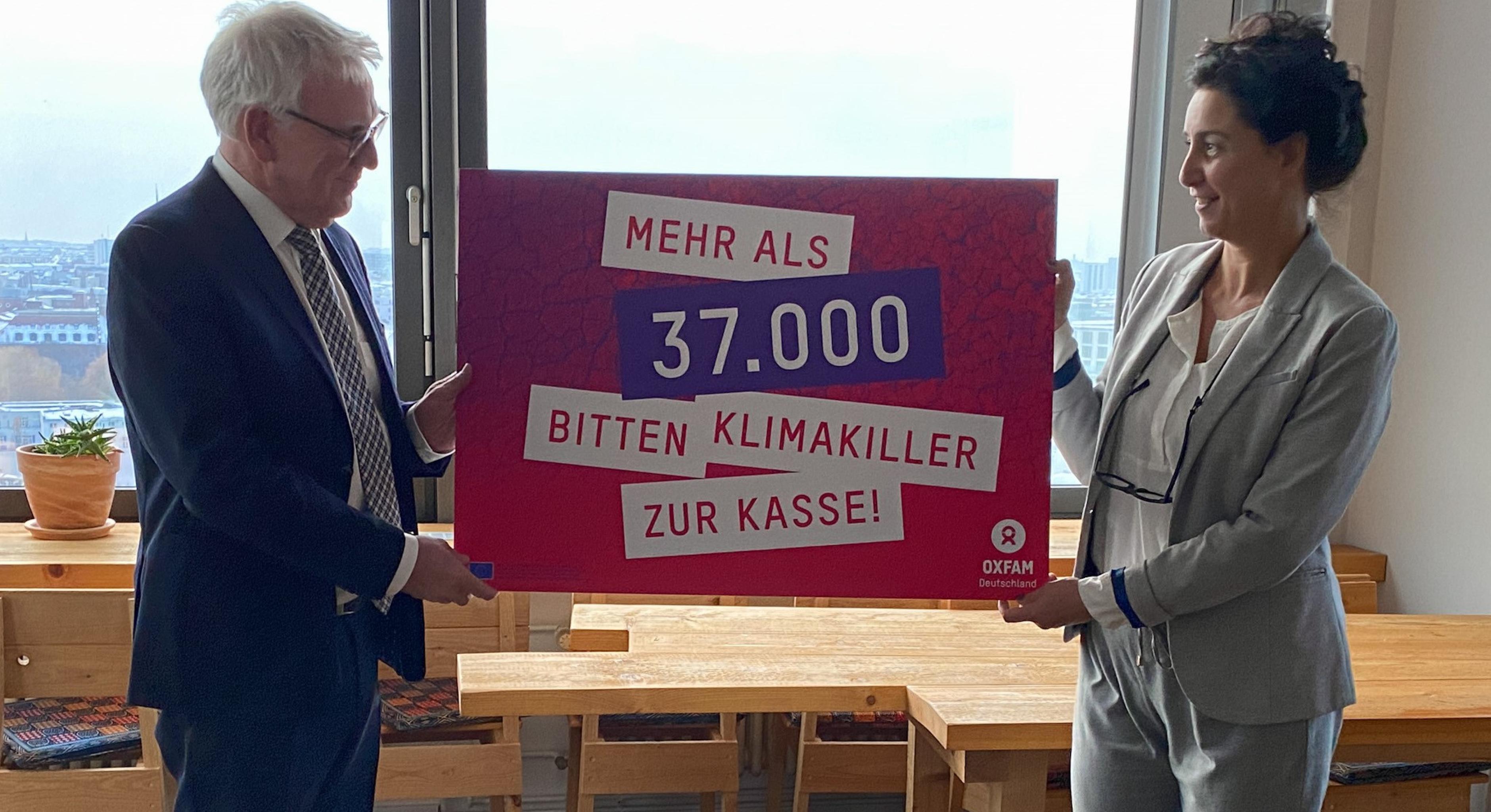 Zwei Menschen halten ein Schild hoch, auf dem steht: Mehr als 37.000 bitten Klimakiller zur Kasse. Dies bezieht sich auf eine Unterschriftenaktion, die Aufmerksamkeit auf eine gerechte Klimapolitik lenken soll.