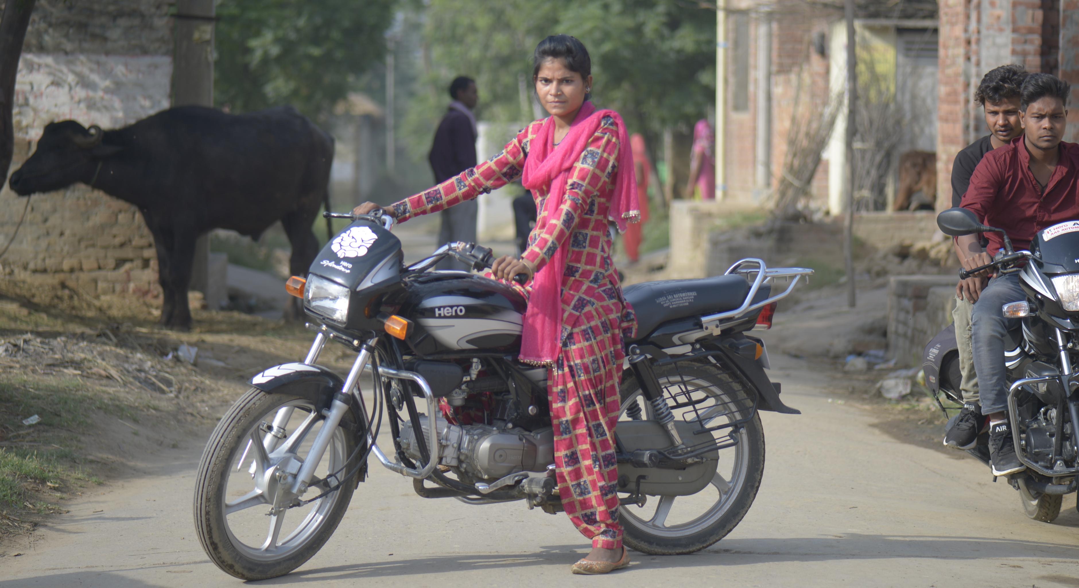 Eine junge indische Frau sitzt auf einem Motorrad; neben ihr zwei Männer auf einem anderen Motorrad