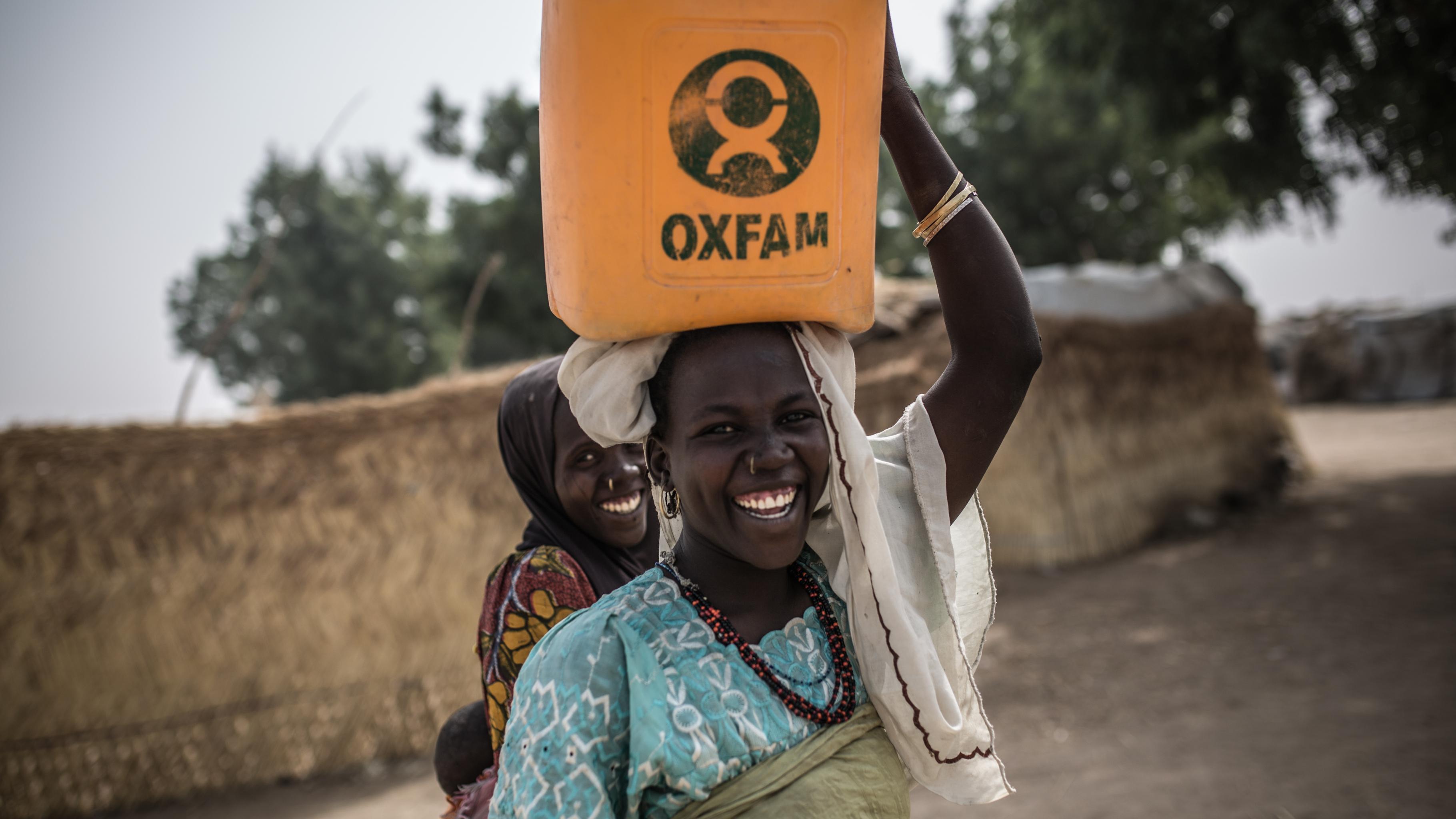 Zwei Frauen lachen; eine von ihnen transportiert einen Oxfam-Wasserkanister auf dem Kopf.