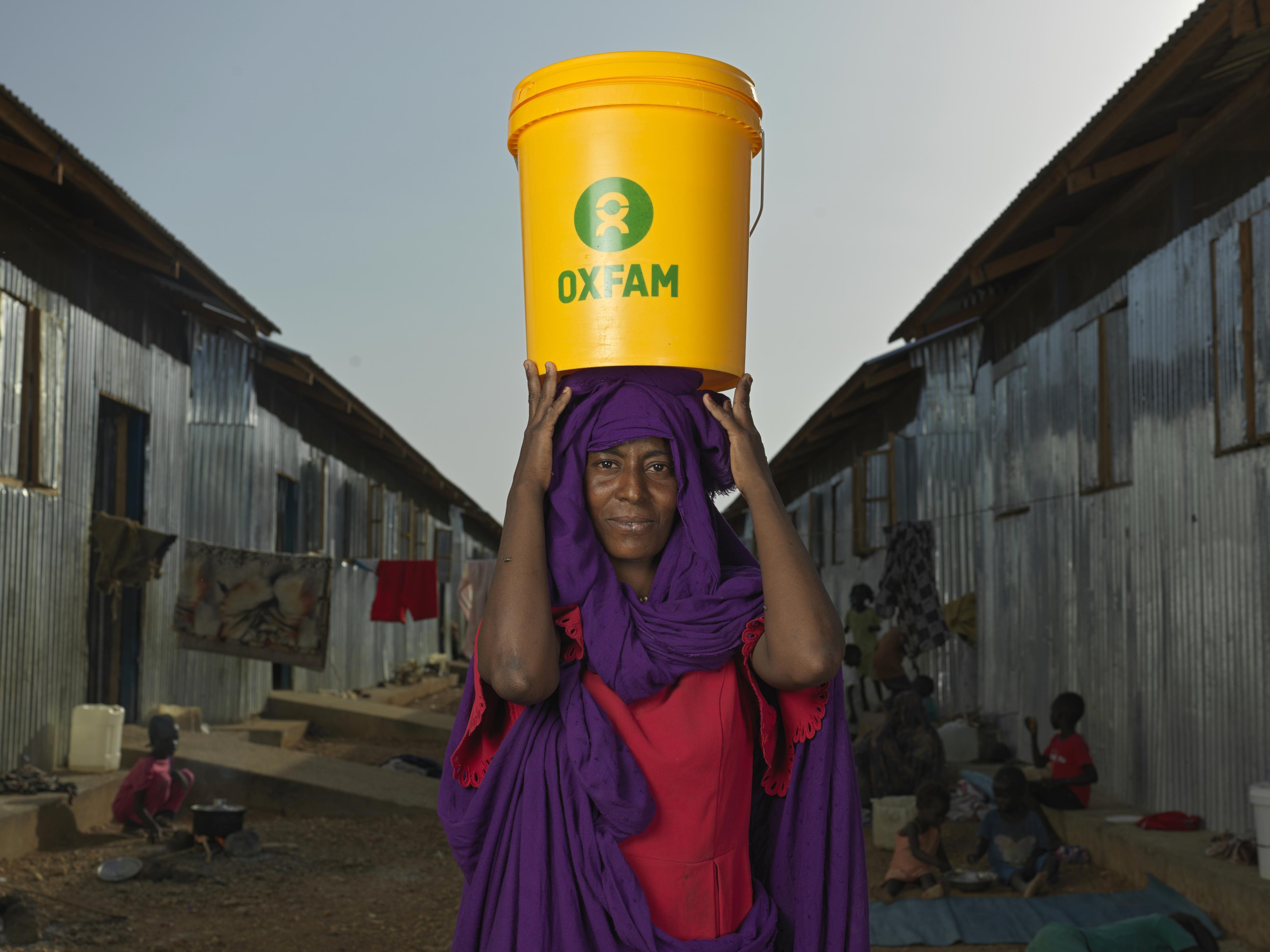Eine Frau scheut in die KAmera. Auf dem Kopf trägt sie einen Wassereimer mit der Aufschrift Oxfam.