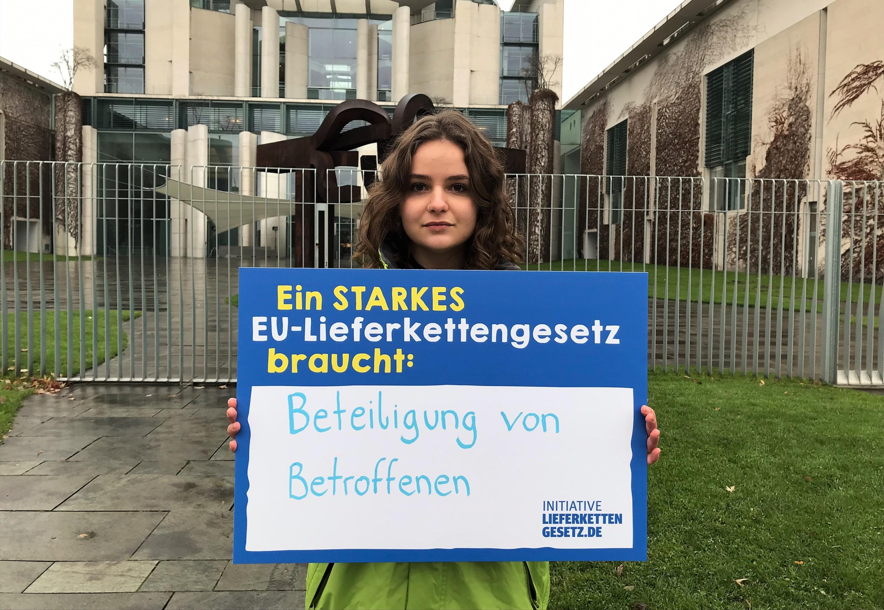 Eine Oxfam-Mitarbeiterin vor dem Kanzleramt mit einem Schild der Initiative Lieferkettengesetz. Aufschrift: "Ein starkes EU-Lieferkettengesetz braucht: Beteiligung von Betroffenen"
