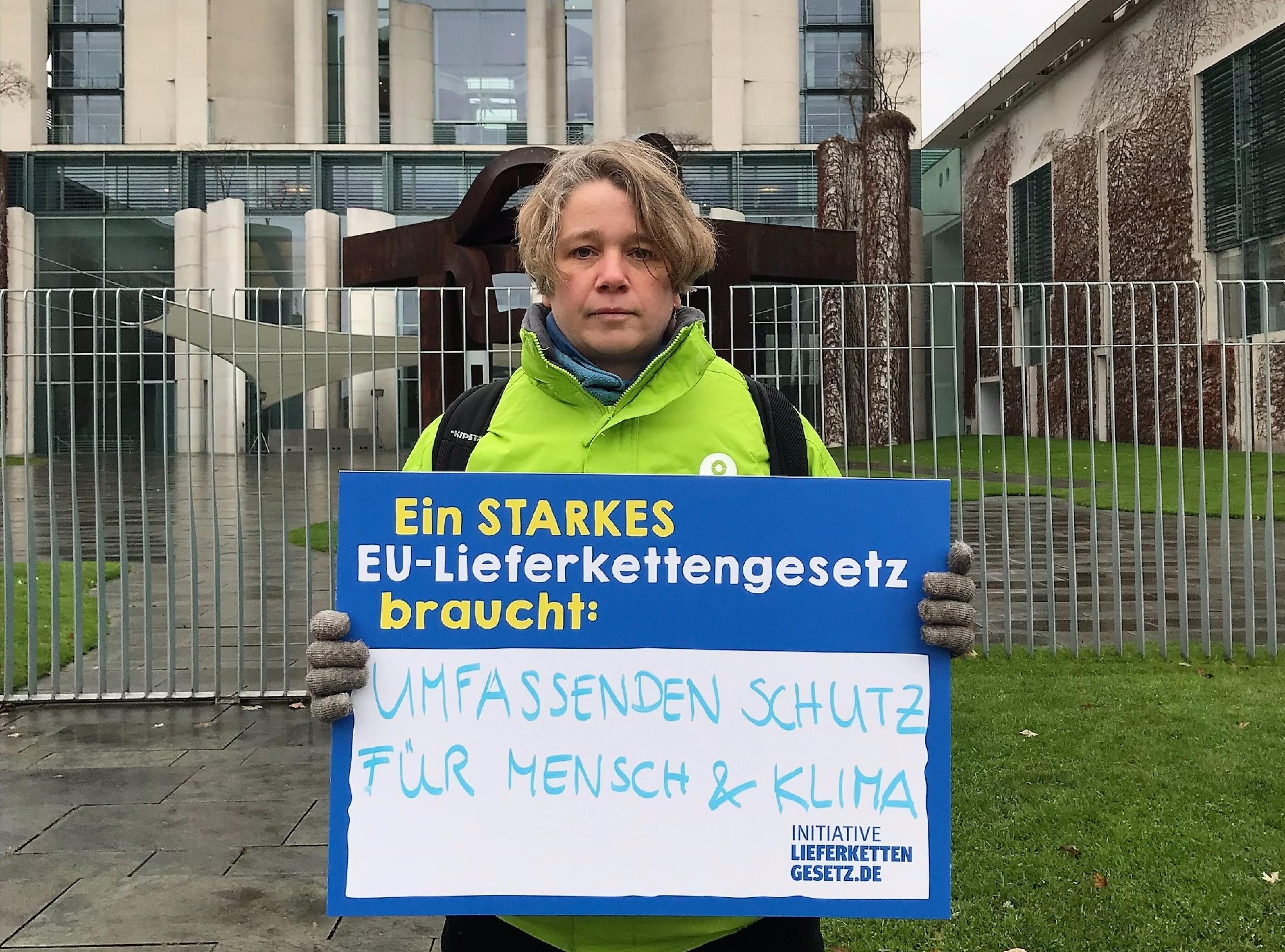 Eine Oxfam-Mitarbeiterin in grüner Oxfam-Jacke steht vor dem Bundeskanzleramt und hält ein Schild der Initiative Lieferkettengesetz vor ihrem Körper. Aufschrift: "Ein starkes EU-Lieferkettengesetz braucht: Umfassenden Schutz für Mensch und Klima".