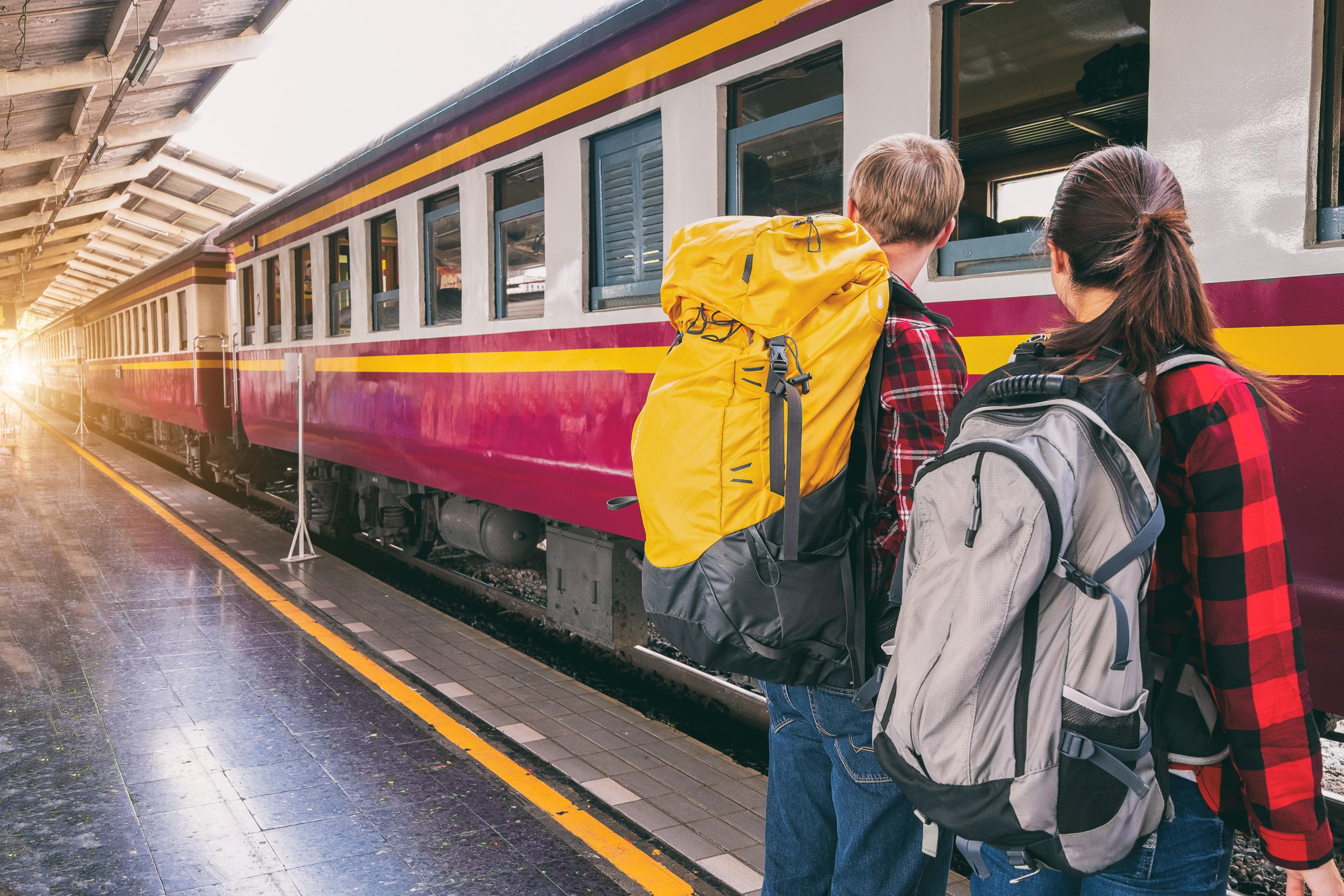 Zwei Personen stehen auf einem Bahnsteig und schauen in Richtung des einfahrenden Zuges. Sie tragen beide Rucksäcke auf ihren Rücken.