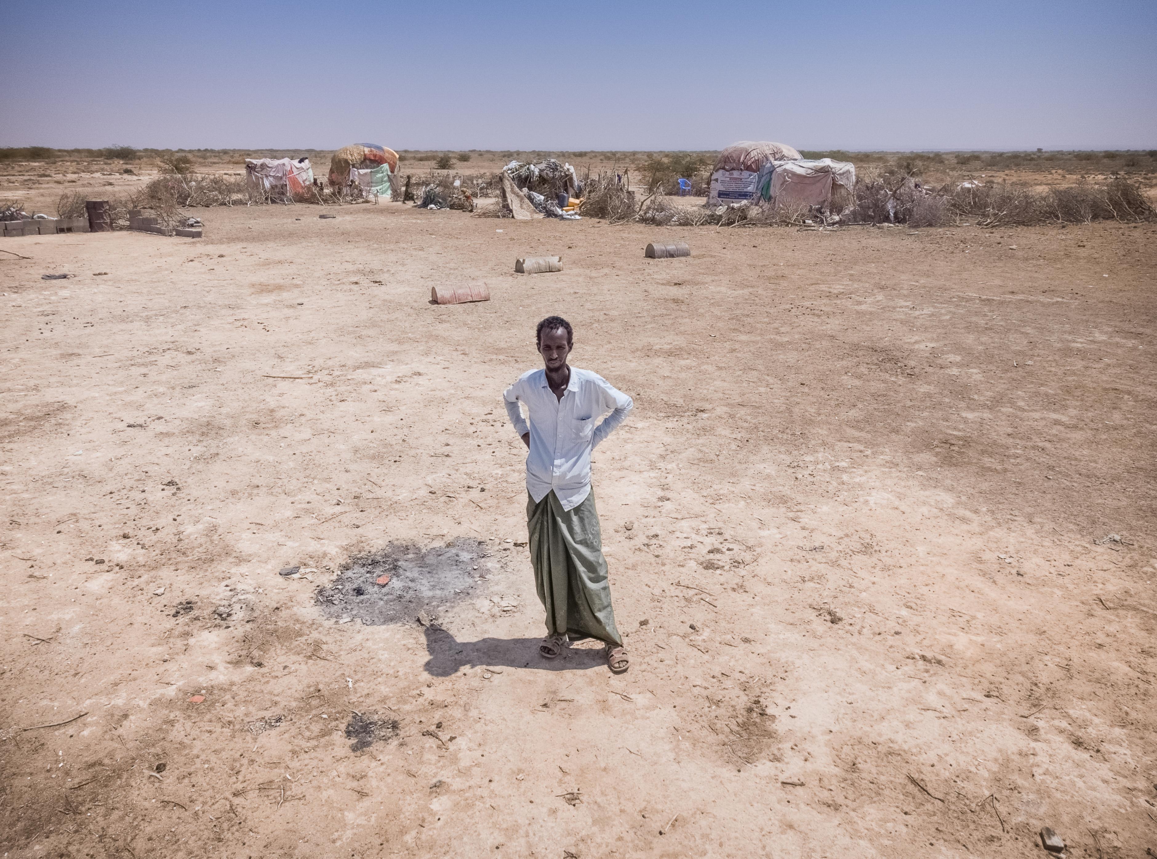 Abdulahi Farah Isse steht, bekleidet mit einem weißen Shirt und einem grauen Wickelrock, auf einem sehr trockenen Boden. Im Hintergrund ist seine Unterkunft zu sehen.