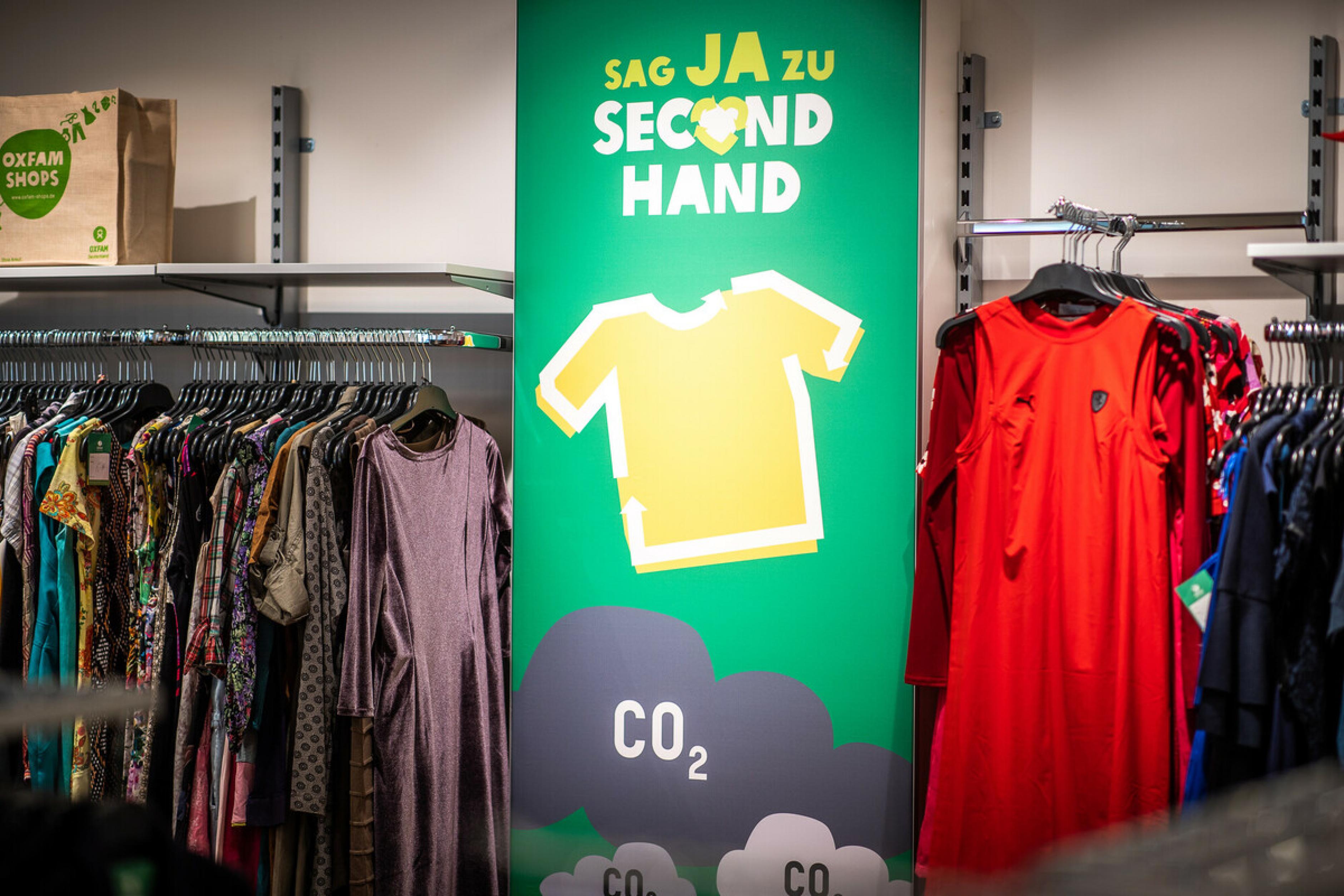 Im Oxfam Shop Berlin-Weißensee hängt Kleidung in bunten Farben auf Kleiderständern. Dazwischen hängt ein Schild mit der Aufschrift „Sag ja zu Second Hand“.