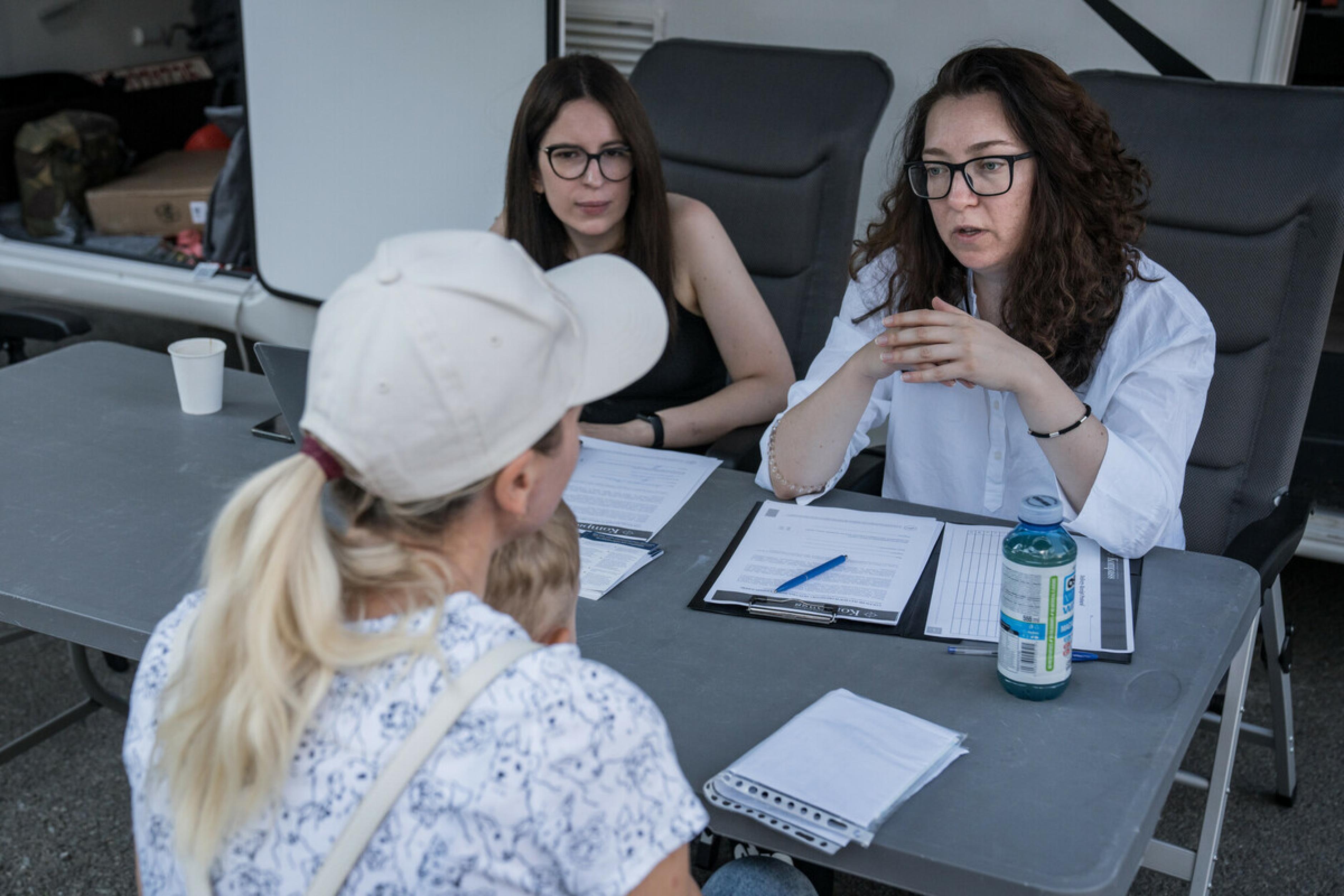 Vor dem Wohnmobil sitzen drei Frauen um einen Tisch und sprechen miteinander. Die eine Frau, eine Geflüchtete, hat ein Kind auf dem Schoß. Die anderen beiden Frauen arbeiten bei eLiberare, einer Organisation, die sich gegen Menschenhandel und sexuelle Ausbeutung Geflüchteter in Rumänien einsetzt.
