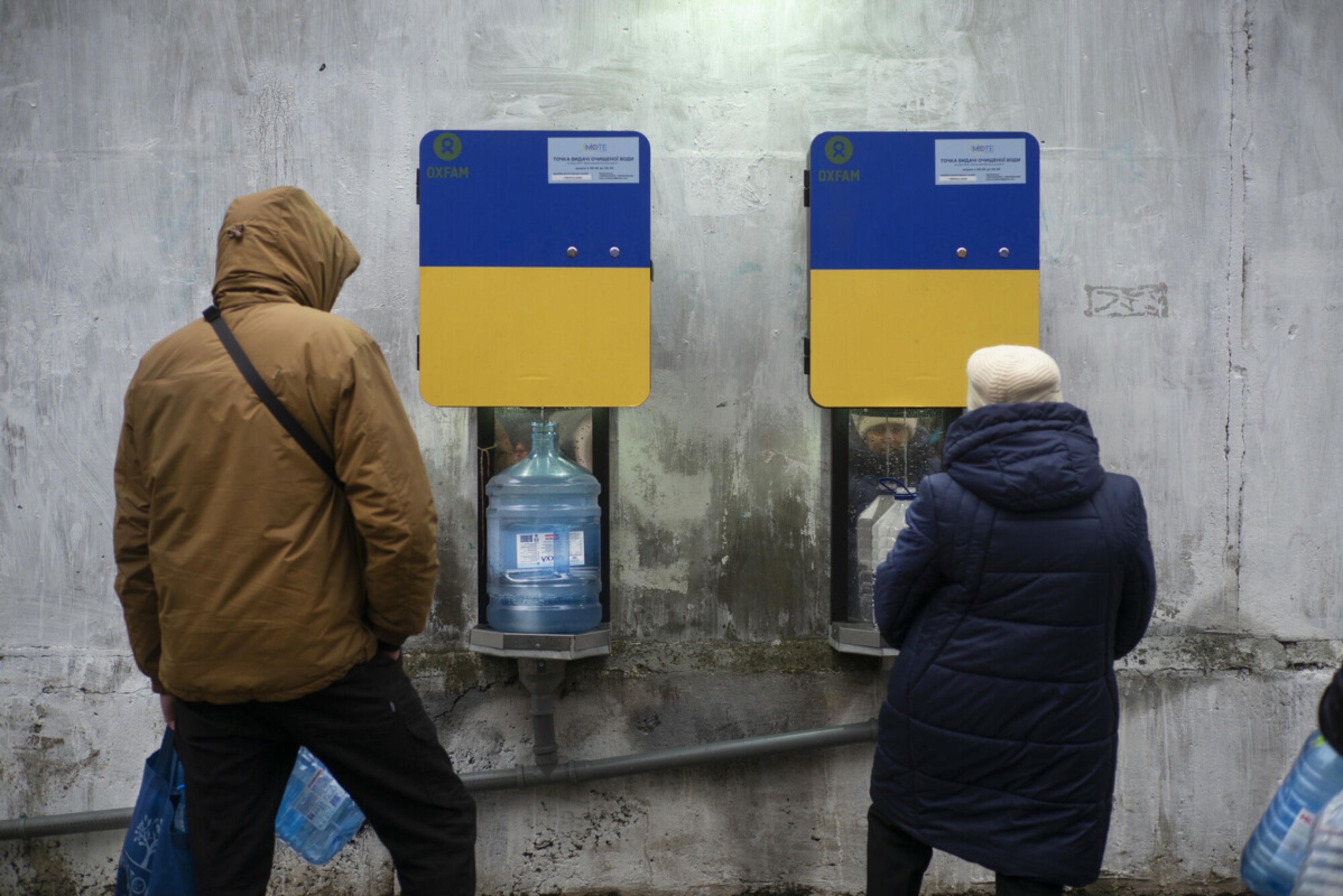 2 Menschen stehen vor zwei Trinkwasserautomaten, aus denen Wasser in ihre Flaschen läuft.