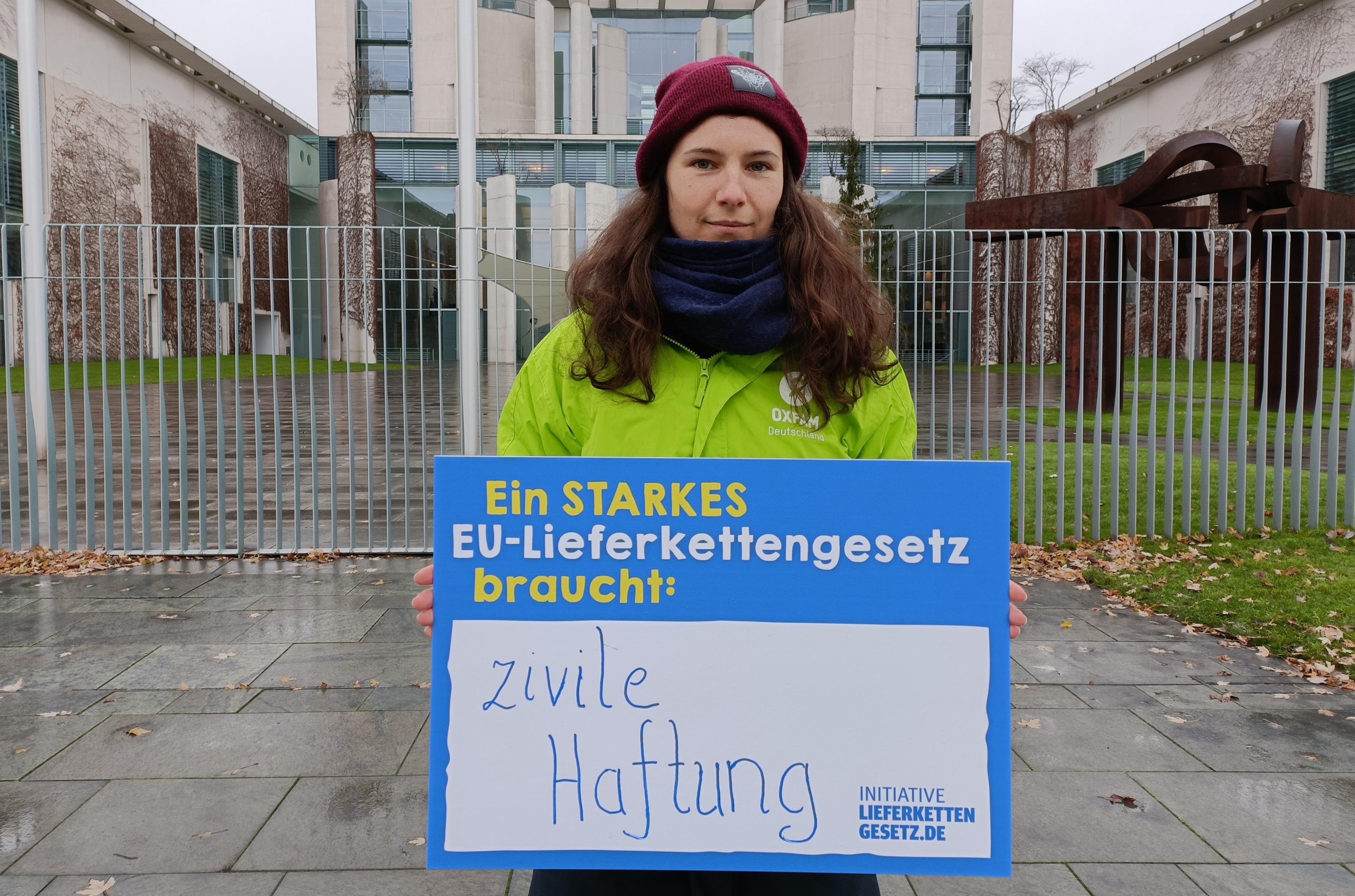 Oxfam-Mitarbeiterin, in grüner Oxfam-Jacke, vor dem Kanzleramt mit einem Schild der Initiative Lieferkettengesetz. Aufschrift: "Ein starkes EU-Lieferkettengesetz braucht: Zivile Haftung"