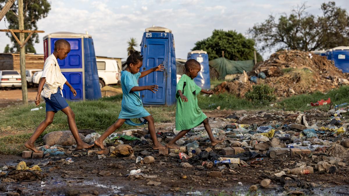 Drei Kinder laufen über einen Weg aus Steinen, Die Umgebung sieht sehr schmutzig aus. Müll liegt auf dem Weg. Abwasser scheint aus nahegelegenen Toiletten auszutreten.