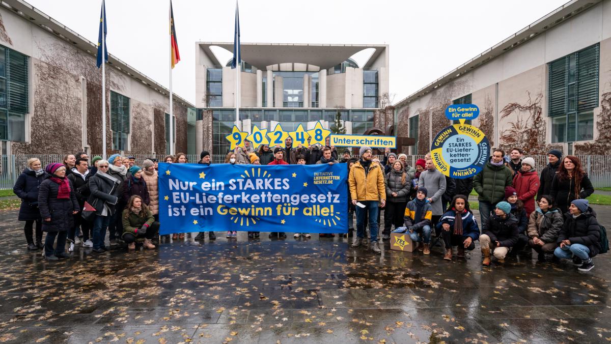 Aktivist*innen der "Initiative Lieferkettengesetzt" mit einem Banner "Nur ein starkes EU-Lieferkettengesetz ist ein Gewinn für alle!" vor dem Bundeskanzleramt. In der hinteren Reihe halten einige Aktivist*innen die Anzahl der eingereichten Unterschriften (90.248) hoch. Rechts ist ein Glücksrad zu sehen, das den "Gewinn für alle" als Hauptgewinn anzeigt. 