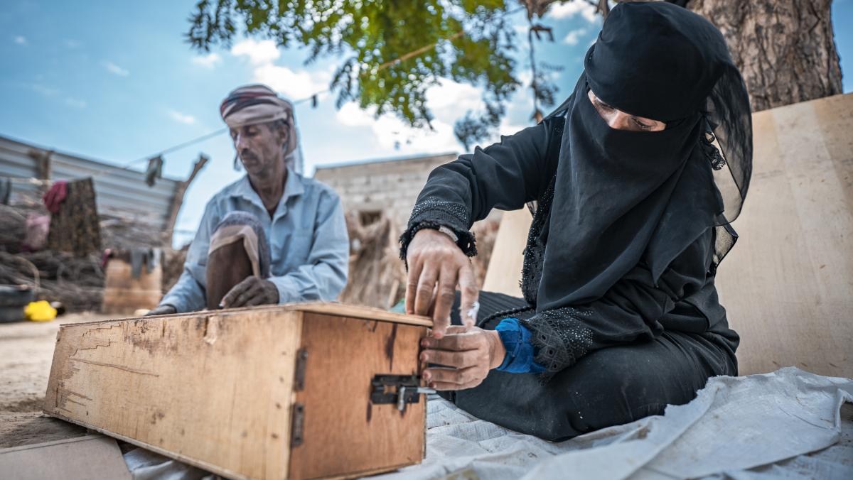 Nazrah (rechts) sitzt zusammen mit ihrem Mann (links) auf einer Decke und baut einen Bienenstock. Sie trägt ein schwarzes Gewand und ist verschleiert. Im Hintergrund sind ein Baum und eine Wäscheleinesichtbar.
