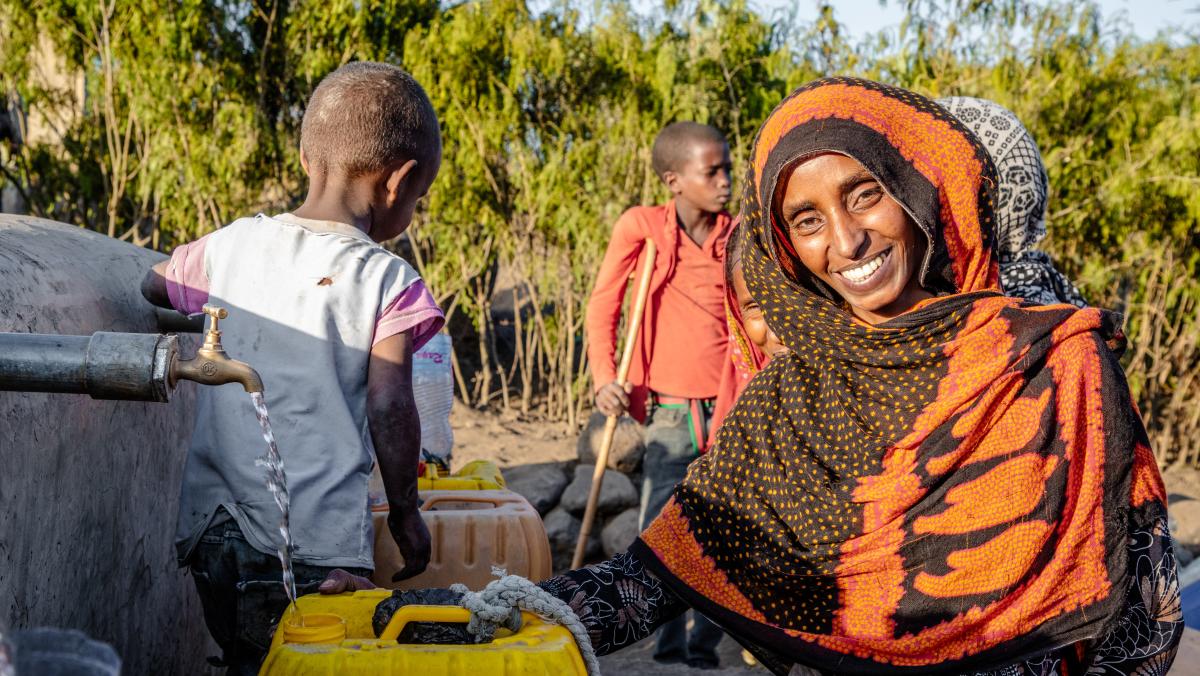 Seadiya Mohamed lächelt in die Kamera, während sie Wasser aus einem laufenden Hahn in ihren Kanister schöpft. Im Hintergrund stehen zwei Kinder.