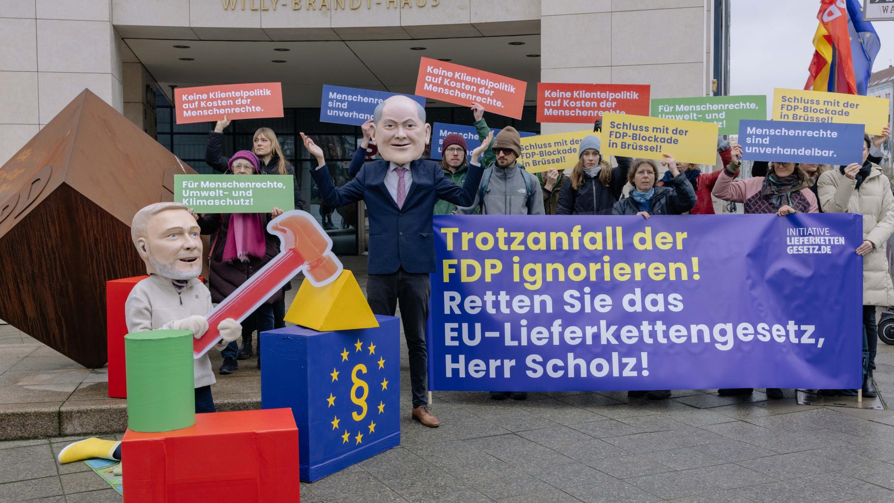    OOxfam Vor dem Willy-Brandt-Haus in Berlin posieren Aktivist*innen mit Plakaten für ein EU-Lieferkettengesetz. Davor schlägt Big-Head-Bundeskanzler Olaf Scholz ratlos die Hände über dem Kopf zusammen, während Big-Head-Finanzminister Christian Lindner wie ein kleines Kind fröhlich mit einem Hammer im EU-Bauklötze-Haus um sich schlägt.