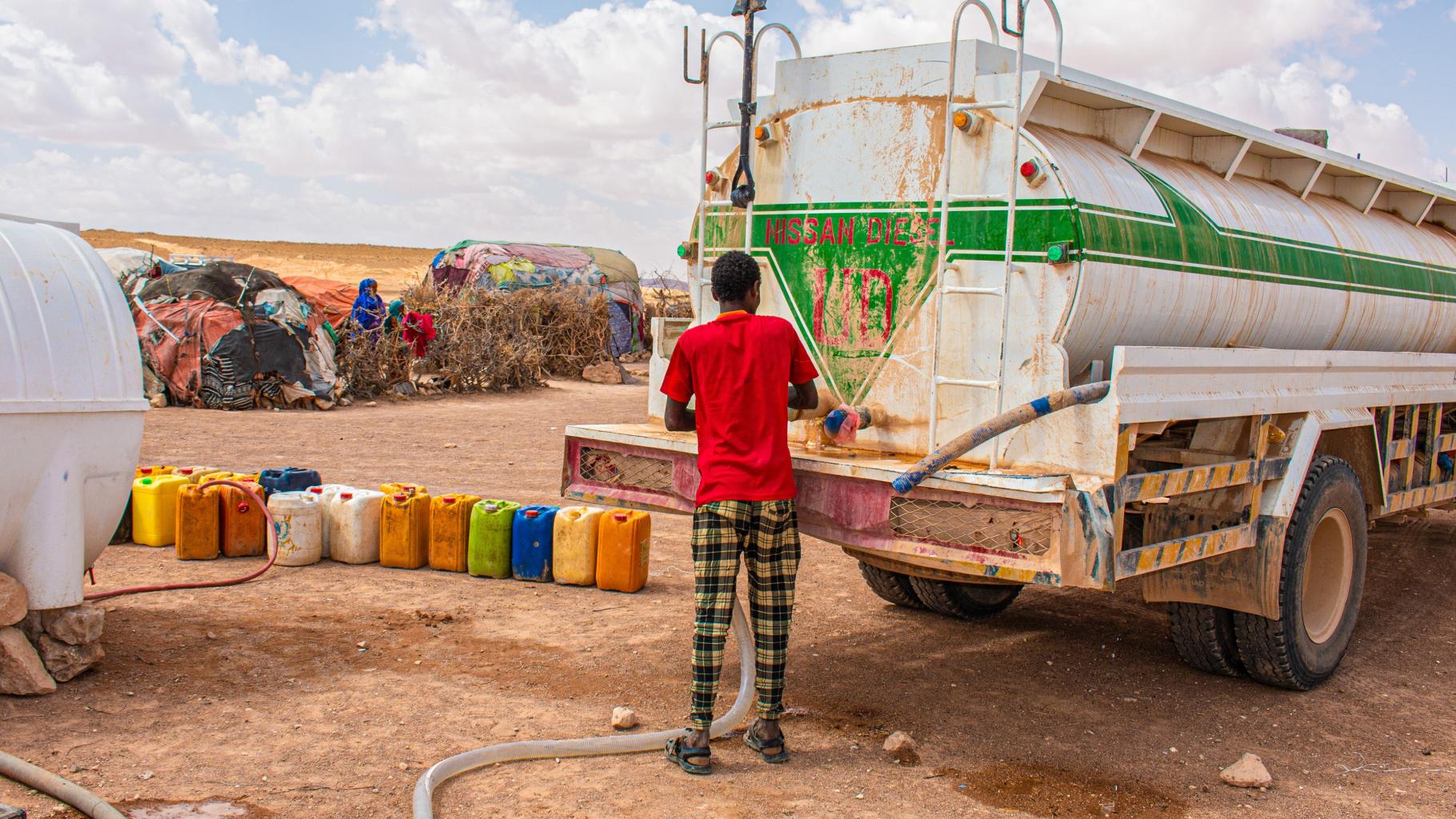 Ein Mann, bekleidet mit einem roten T-Shirt und einer gelb-schwarz karierten Hose, verbindet einen großen Schlauch mit einem LKW voll sauberem Trinkwasser