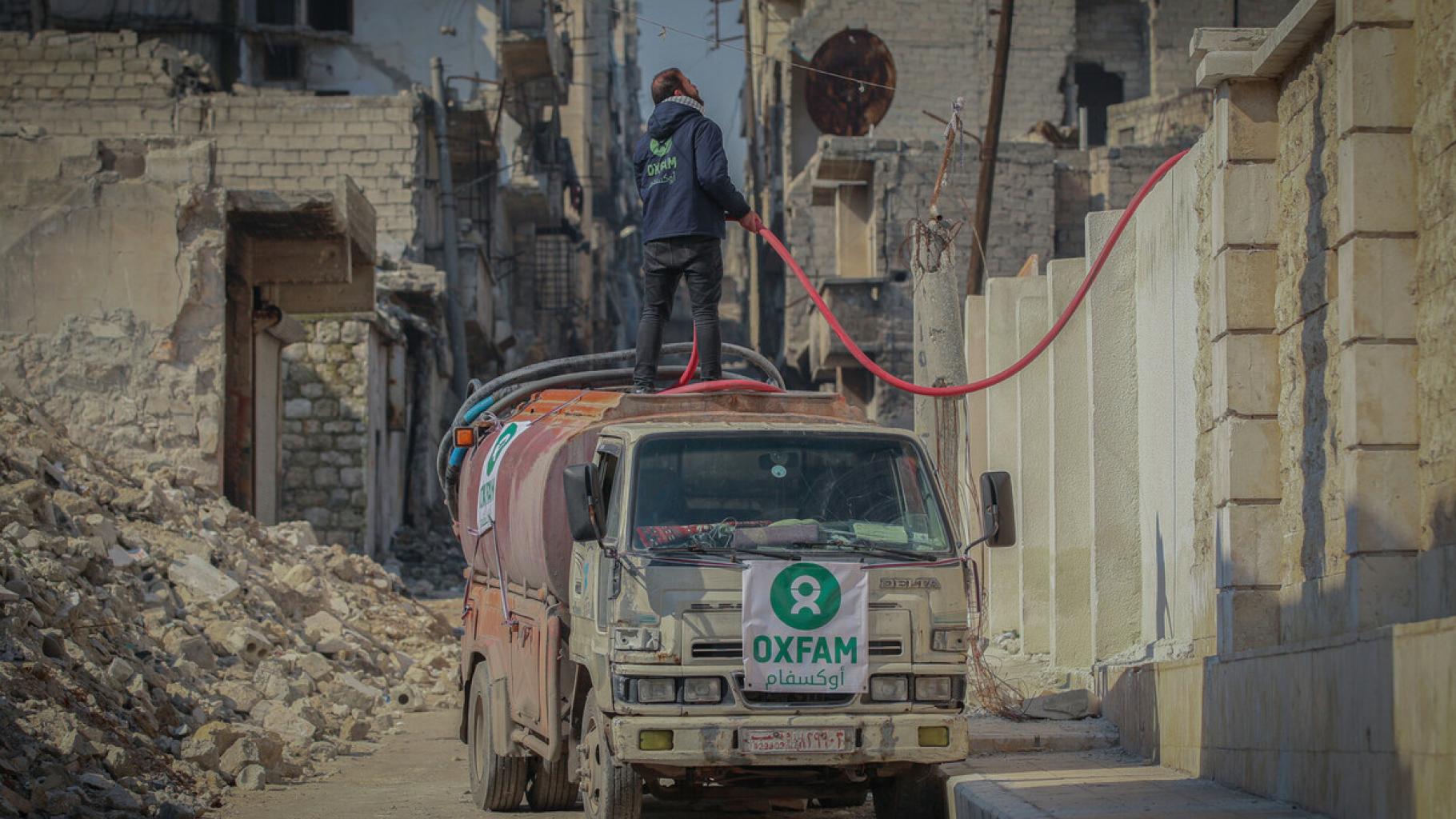 Ein Tanklastwagen in einer Straße in Aleppo. Auf dem Wagen steht ein Mann mit einem Schlauch in der Hand. Links neben dem Wagen sieht man die Ruinen eines Hauses.