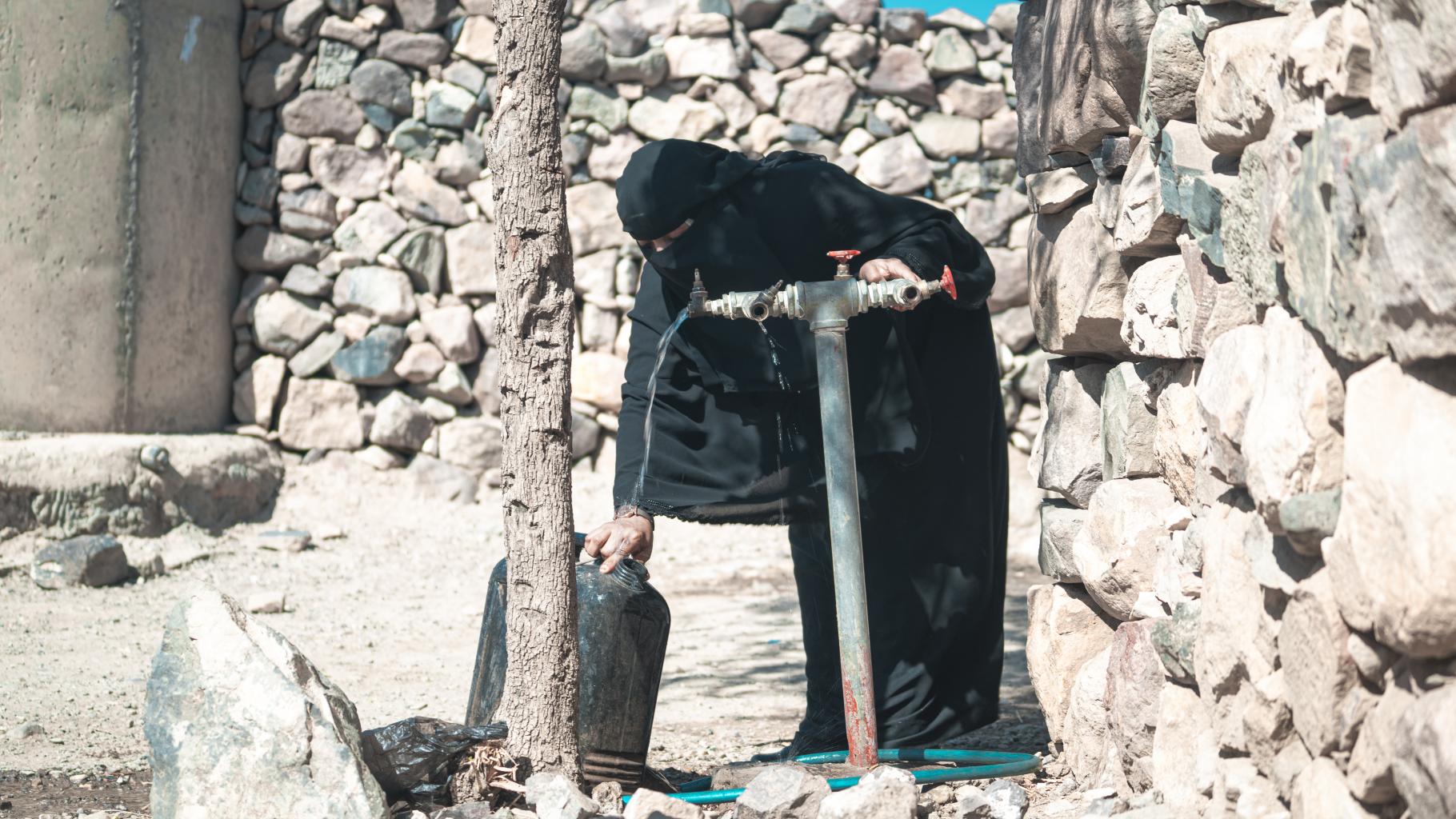 Haneen* (in schwarzem Gewand und mit Niqab bekleidet) dreht einen Wasserhahn vor ihrem Haus auf, um einen Wasserkanister zu befüllen.