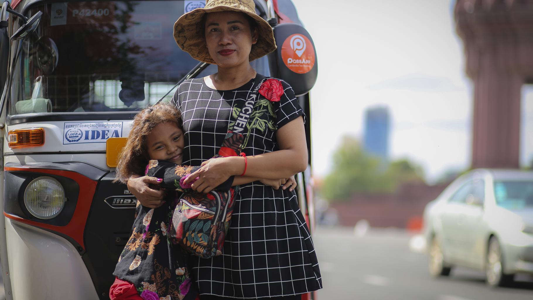 Eine Frau mit Hut steht vor ihrem Tuk-Tuk und hält ihre Tochter im Arm.
