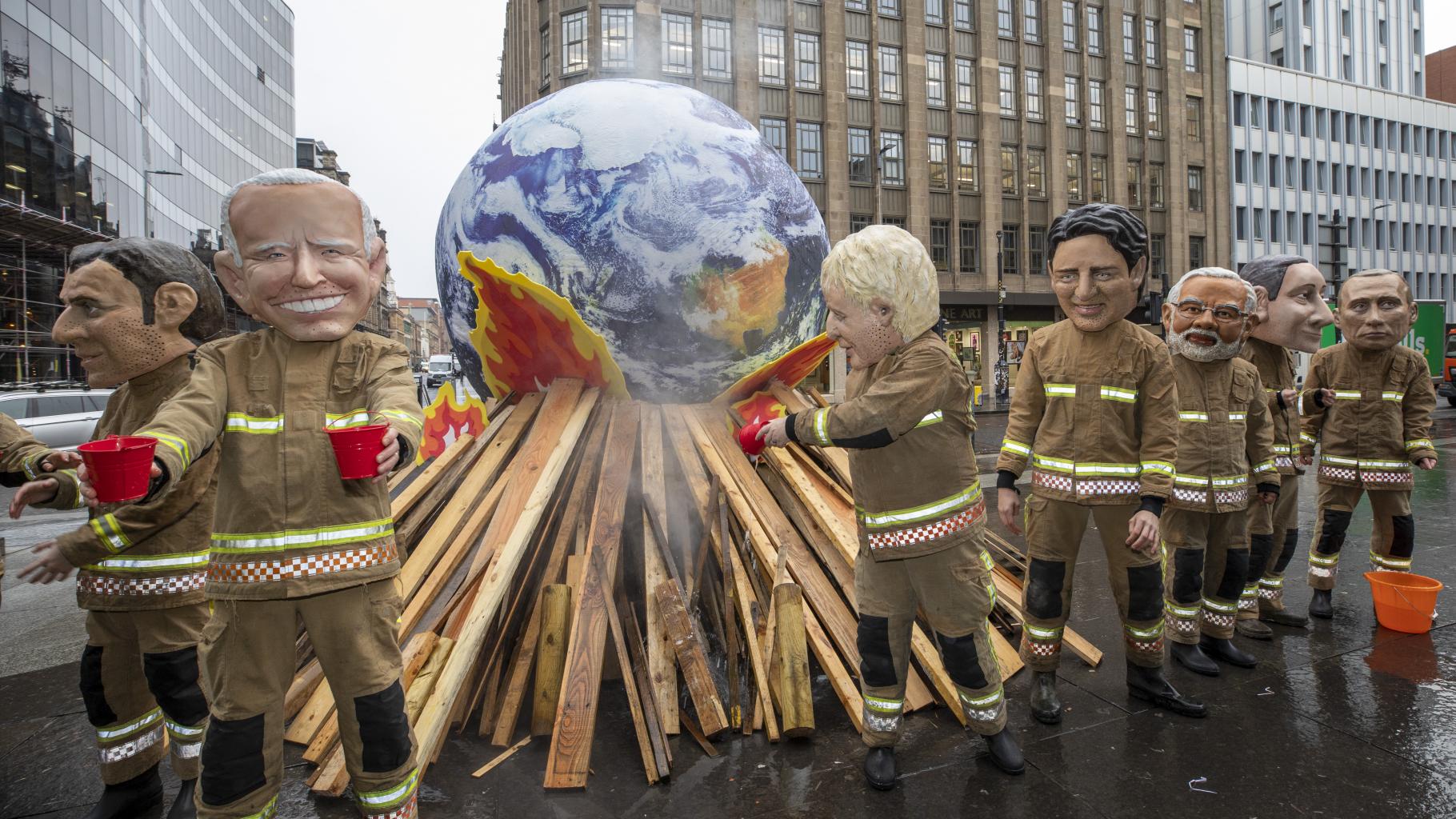 Oxfam-Aktivist*innen posieren mit überdemensionalen Pappmasche-Köpfen der mächtigsten Regierungspersonen und als Feuerwehrmenschen verkleidet bei der COP26 in Glasgow. Sie stehen vor einer brennenden Erdkugel und versuchen sie erfolglos zu löschen.