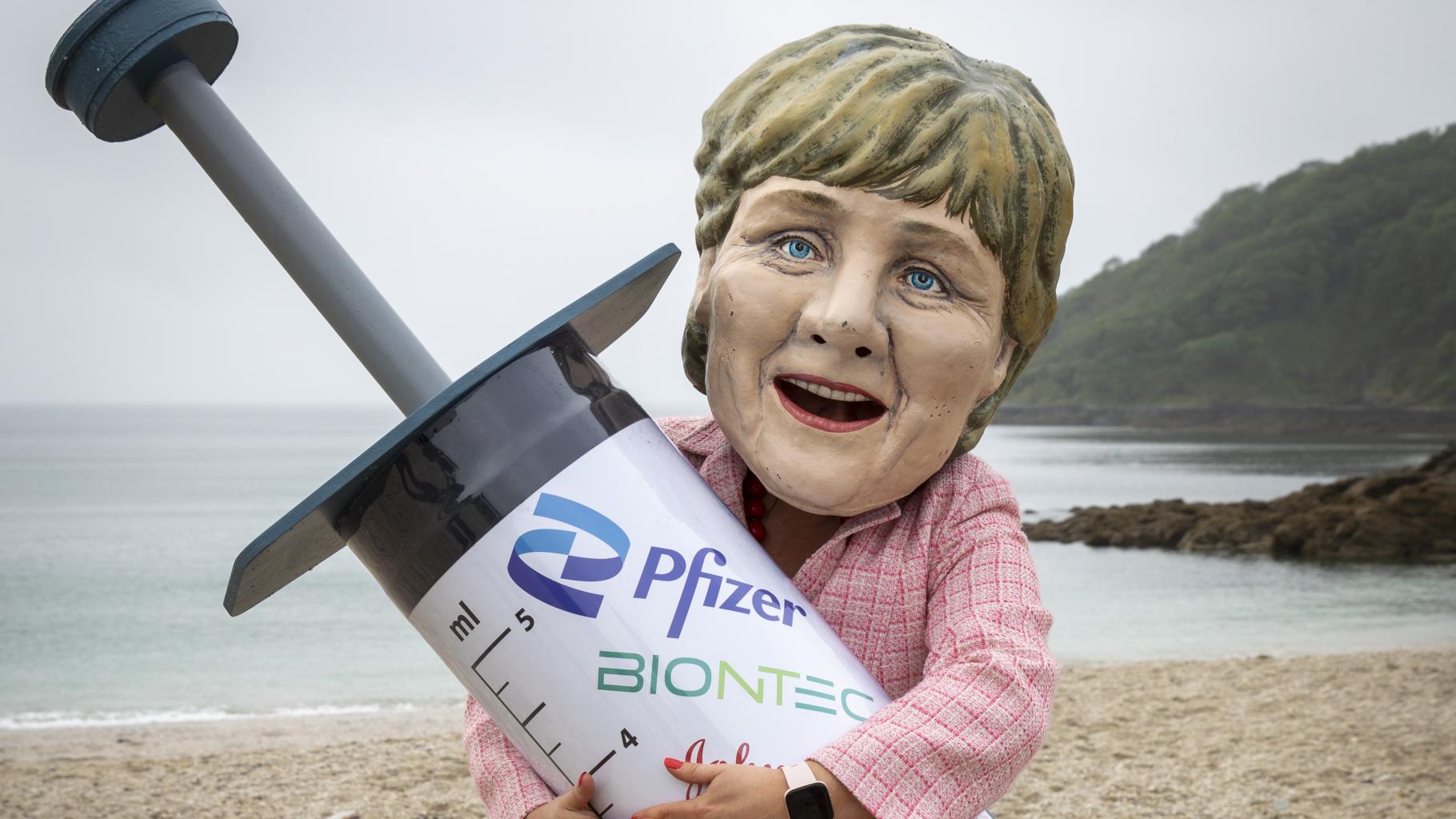 Ein*e Aktivist*in ist mit einem riesigen Angela-Merkel-Kopf bekleidet hält eine riesige Spritzen-Atrappe im Arm. Im Hintergrund sind Strand und Meer zu sehen.
