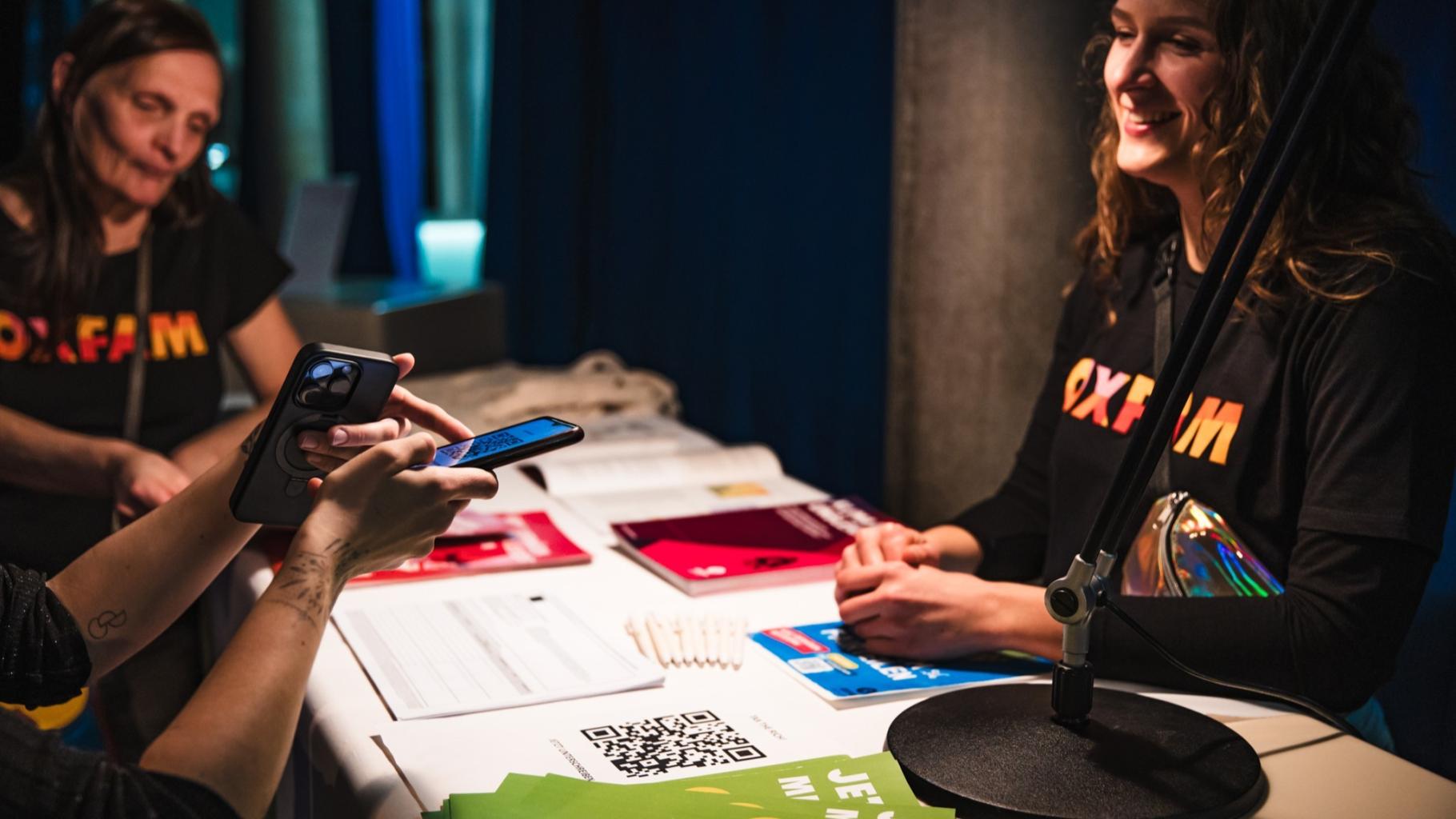 Zwei Oxfam-Mitarbeitende in schwarzen T-Shirts mit buntem Oxfam-Logo stehen um einen Info-Tisch und sprechen mit einer Person, die Infos zu unserer Petition erhalten will. Von dieser Person sind in der linken Ecke des Fotos nur die Arme und Hände sichtbar, sie macht mit ihrem Handy ein Foto von einem QR-Code.