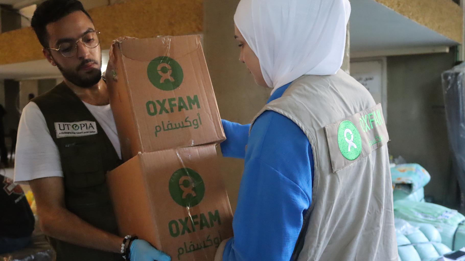 Eine Person übergibt zwei Kartons mit Oxfam-Aufschrift an eine zweite Person. Im Hintergrund stehen Feldbetten.
