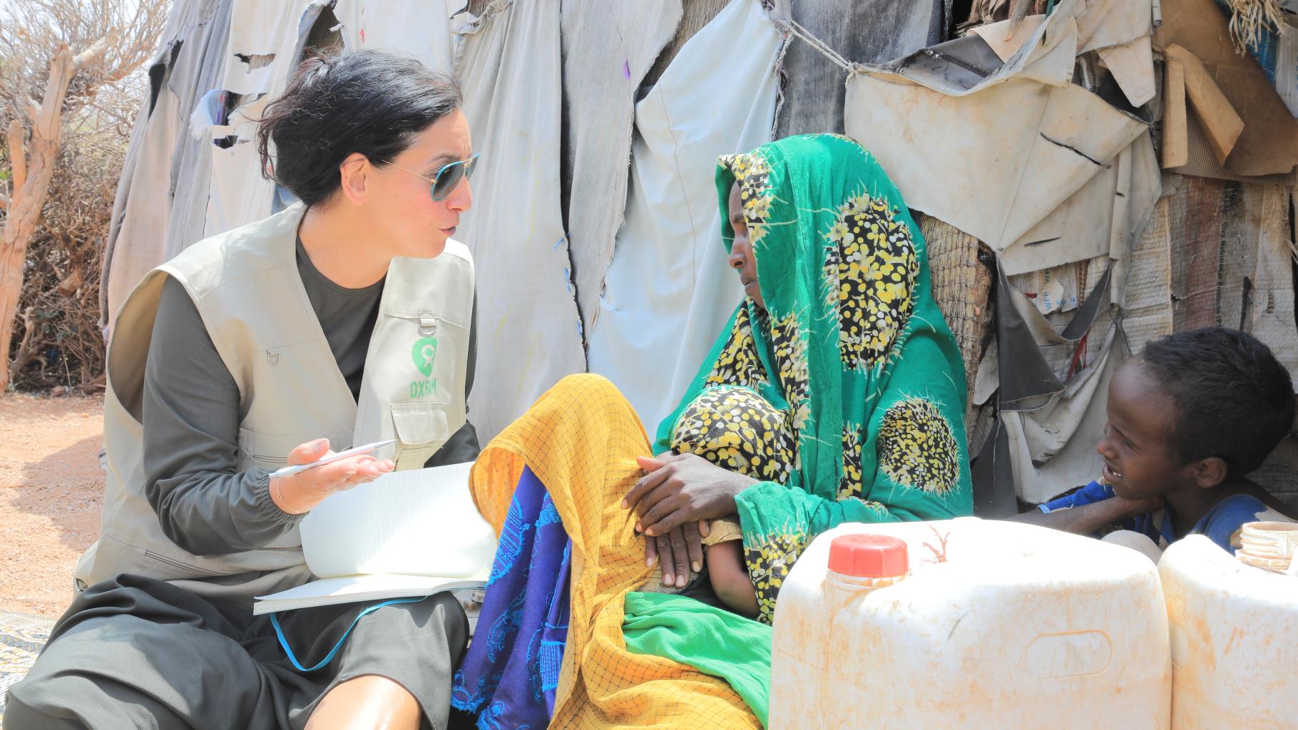 Searp Altinisik und Anisa Jama Hassan sitzen sich gegenüber und sprechen miteinander. Im Hintergrund ist das Geflüchtetencamp sichtbar. Neben Anisa Jama Hassan sitzt eins ihrer Kinder.