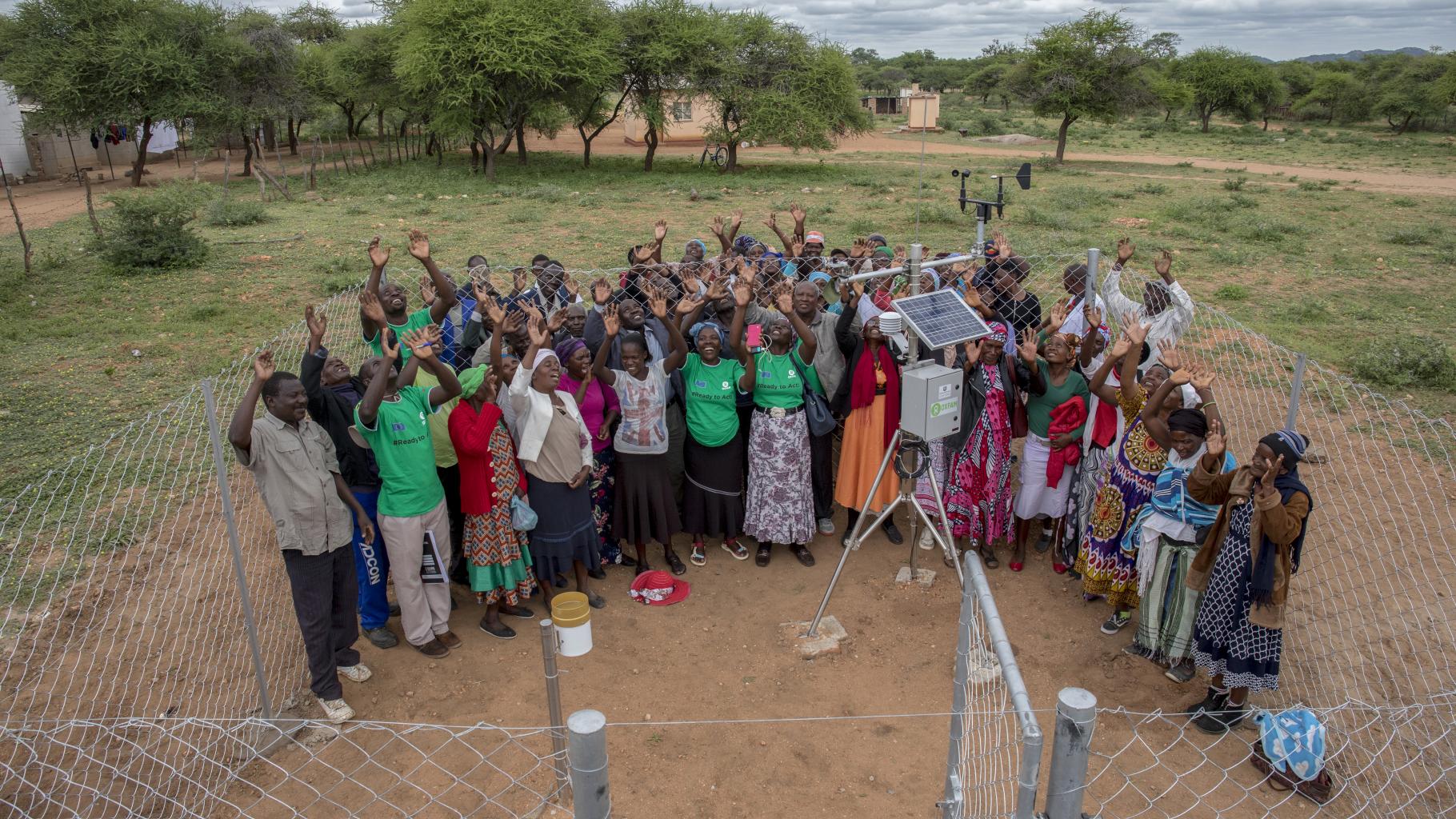 Eine Gruppe von Menschen steht bei einer Wetterstation in einer ländlichen Region in Simbabwe. Sie strecken freudig ihre Arme in die Luft.