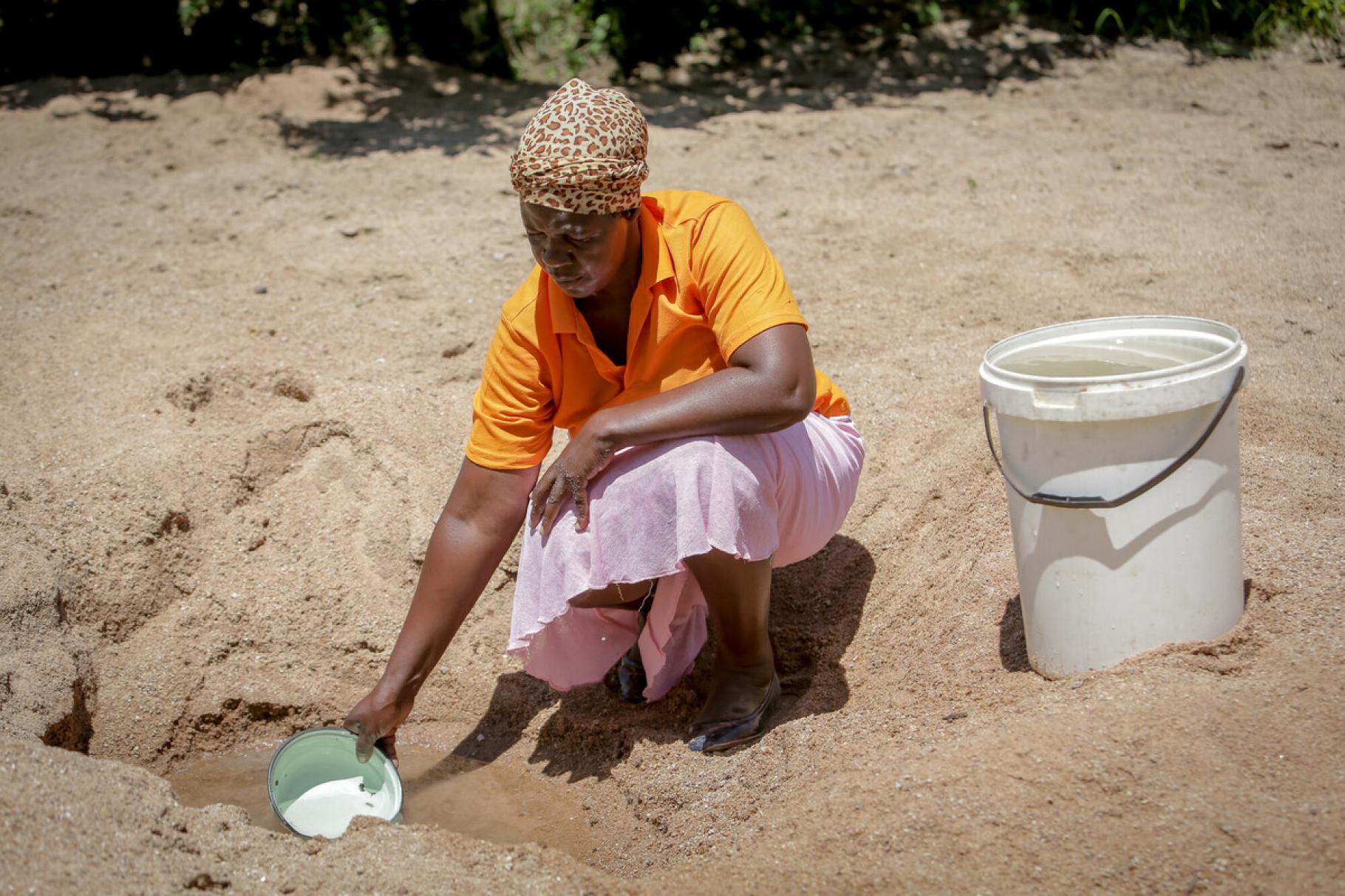 Eine Frau hockt im Sand, vor ihr ein selbst gegrabenes Wasserloch im Boden. Sie schöpft daraus Wasser mit einer kleinen Schüssel. Neben ihr steht ein großer Eimer, der bereits mit Wasser gefüllt ist.