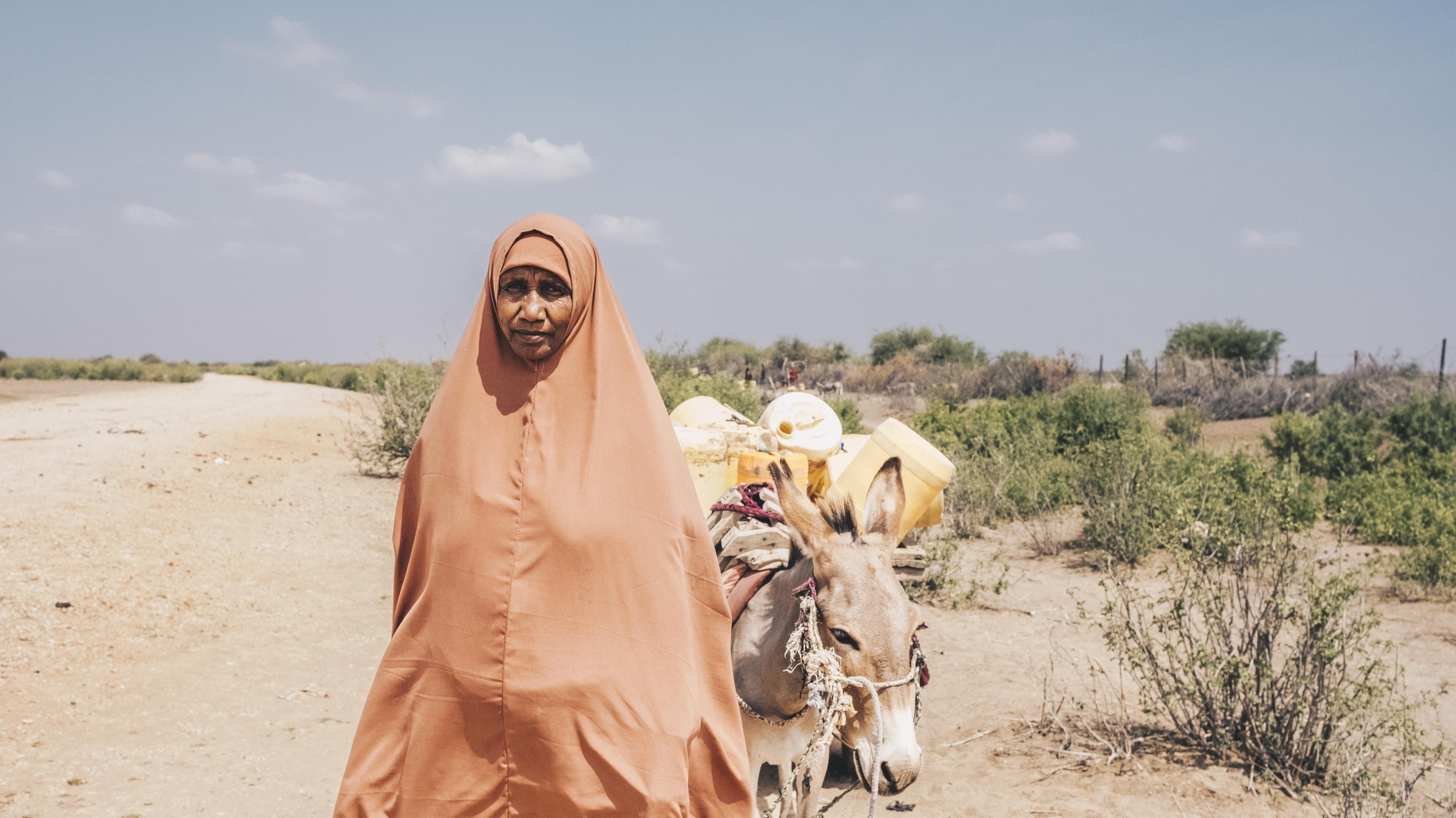 Eine Frau mit einem Esel in einer trockenen, sandigen Gegend. Sie blickt ernst in die Kamera. Die Sonne scheint. Die Wasserkanister auf dem Esel sehen leer aus.