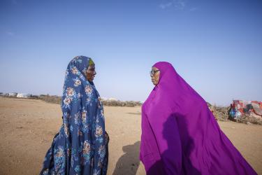Idabo trägt ein lila Kleid und unterhält sich mit einer Person (blaues Kleid). Im Hintergrund ist eine trockene Landschaft und Teile des Camps zu sehen.