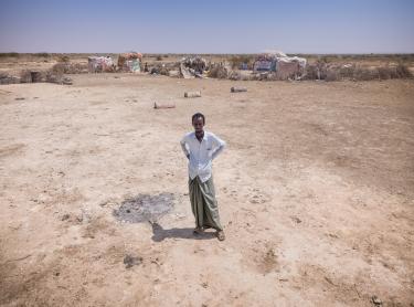 Abdulahi Farah Isse steht, bekleidet mit einem weißen Shirt und einem grauen Wickelrock, auf einem sehr trockenen Boden. Im Hintergrund ist seine Unterkunft zu sehen.