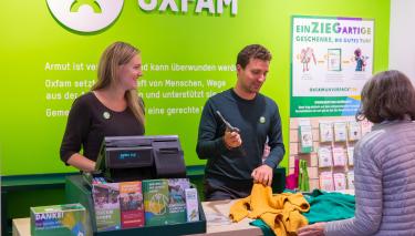 Zwei Ehrenamtliche stehen an einer Kasse in einem Oxfam-Shop und kassieren die Kleidung einer Kundin ab