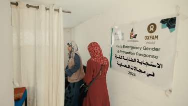 Zwei Frauen betreten einen durch einen weißen Vorhang geschützten Raum. An der Wand ist ein Oxfam-Poster mit der Aufschrift für humanitäre Hilfe zu sehen.