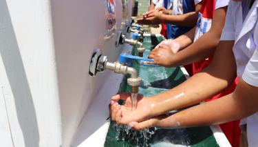 Grundschulkinder stehen nebeneinander an einem länglich gezogenen Wasserhahn und waschen sich die Hände.