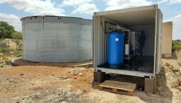 In einem Metallcontainer steht eine elektrische Anlage. Dahinter steht ein großer Wassertank aus Metall.