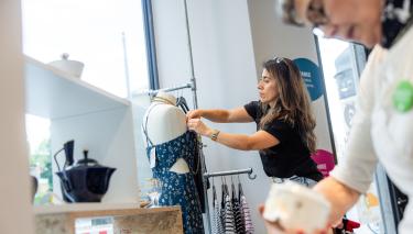 Eine Frau zieht einer Schaufensterpuppe ein Kleid an. Die zweite Frau schaut ein Stück Geschirr an.