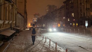 Eine verschneite Straße in Kyiv. Es ist dunkel. Auf dem Gehweg ein Mensch mit Rucksack, auf der Straße die Scheinwerfer eines Autos.