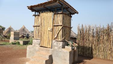 Eine Kabine aus Bambus mit einer Tür und einem Dach aus Bambus. Die Kabine steht auf einem Podest aus Beton.