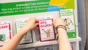Zwei Hände fassen eine Glückwunschkarte mit der Aufschrift „Viel Glück“ in einem Regal. Drumherum stehen viele weitere Grußkarten.