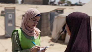 Eine Oxfam Mitarbeiterin spricht mit den Menschen vor Ort über ihre Bedürfnisse Zwei Personen stehen sich gegenüber. Die eine Person notiert etwas in einem Schreibblock.