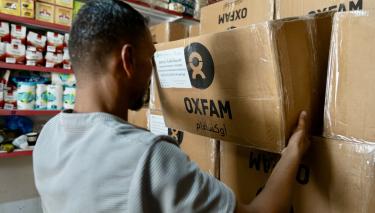 Ein Mann hält einen Karton mit Oxfam-Logo. Im Hintergrund sind Regale mit Nahrungsmitteln zu sehen.