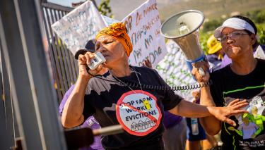 Aktivistin Bettie Fortuin leitet einen Streik vor der Matroosberg Farm in De Doorns, Südafrika. Auf der rechten Seite mit Megafon: Magrieta Prins. Die Gruppe fordert eine regelmäßige und pünktliche Bezahlung der Arbeiter*innen auf der Farm. Eine Frau spricht in ein Megafon, die andere hält das Megafon. Aufschrift auf einem T-Shirt: „Stop Farm Worker Evictions“ „(Stoppt die Zwangsräumung von Farmarbeiter*innen“)