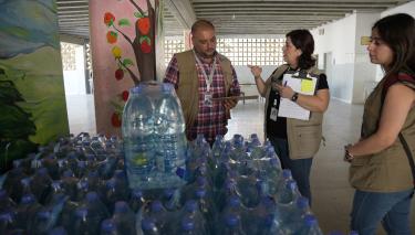 Drei Personen stehen hinter einem großen Stapel Wasserflaschen. Sie halten Klemmbretter in der Hand und sprechen miteinander.