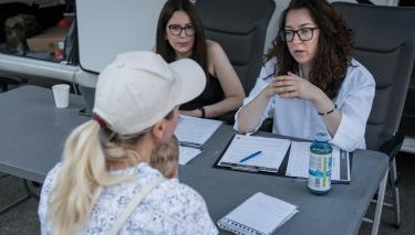 Vor dem Wohnmobil sitzen drei Frauen um einen Tisch und sprechen miteinander. Die eine Frau, eine Geflüchtete, hat ein Kind auf dem Schoß. Die anderen beiden Frauen arbeiten bei eLiberare, einer Organisation, die sich gegen Menschenhandel und sexuelle Ausbeutung Geflüchteter in Rumänien einsetzt.