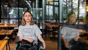Eine junge Frau im Rollstuhl schaut selbstbewusst in die Kamera. Im Hintergrund stehen Tische und Stühle eines industriell eingerichteten Lokals.tergrund 