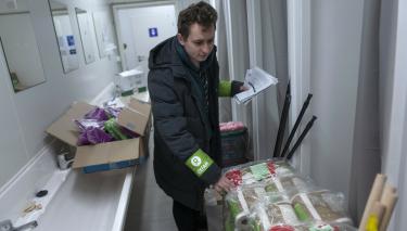 Vladislav Kubrak steht in einem Waschraum vor einem großen Paket Hygienepapierrollen. Hinter ihm befinden sich weitere Kartons mit Hygieneartikeln.