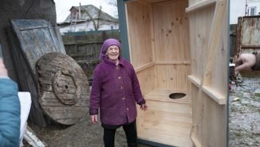Nadiya Pavlenko steht vor ihrer neuen Latrine aus Holz. Sie strahlt in die Kamera.